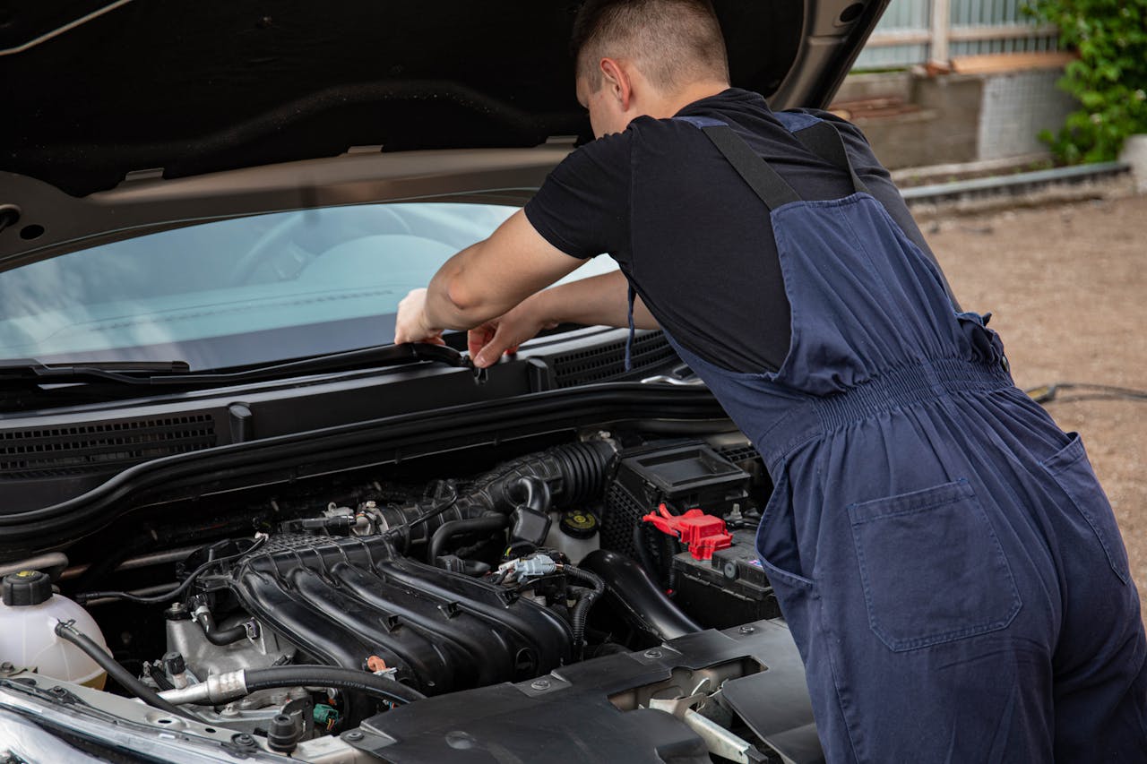 A mechanic in blue overalls works on a car engine outdoors, showcasing vehicle maintenance skills.